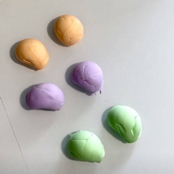 Six morceaux de pâte pastel de taille identique alignés sur une surface : deux orange, deux verts et deux violets, prêts à être modelés en coques de macarons.