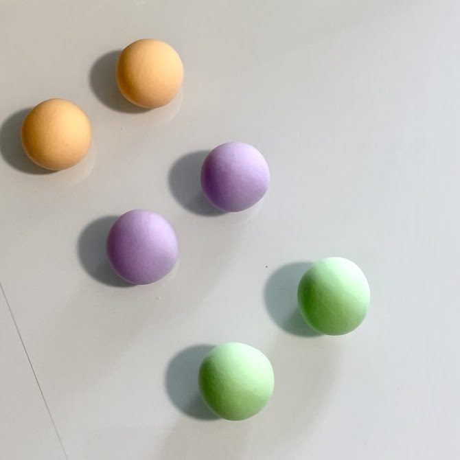 Six boules de pâte pastel parfaitement rondes et lisses posées sur la table : deux orange, deux vertes et deux violettes, prêtes à être transformées en coques de macarons.