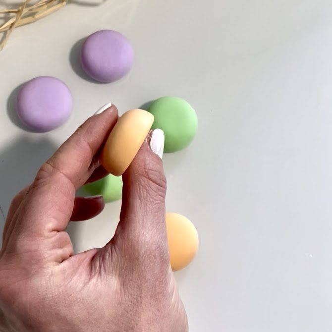 Mains tenant une coque de macaron orange pastel bien formée, vue de profil, avec une surface supérieure arrondie et une base bien plate.