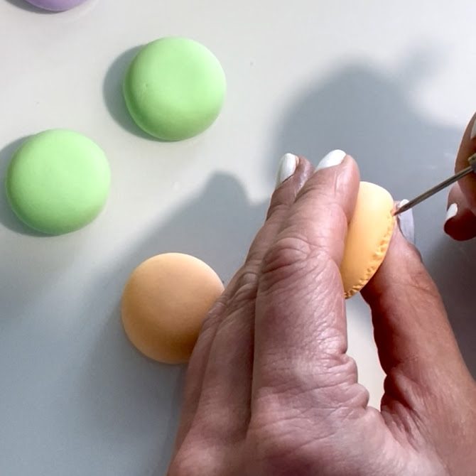 Mains créant de petits points sur la base d'une coque de macaron en foam clay à l'aide d'un outil à pointe fine, pour imiter la texture caractéristique des vraies coques.