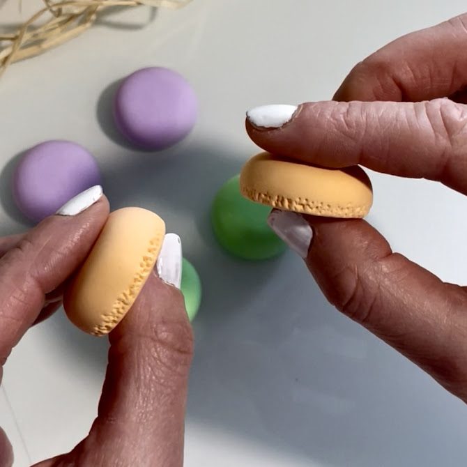Deux mains présentent côte à côte deux coques de macarons en foam clay, vues de profil pour mettre en valeur les petits points texturés soigneusement réalisés à la base.