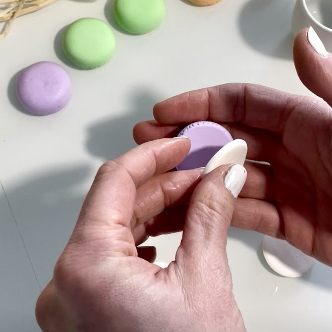 Mains plaçant délicatement un cercle de silk clay sur la base d’une coque de macaron violette pour assembler les deux parties.