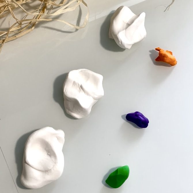 Morceaux de pâte Silk Clay blanche disposés sur une table, accompagnés de petites quantités de Foam Clay violette, orange et verte, prêts à être mélangés pour créer des couleurs pastel.