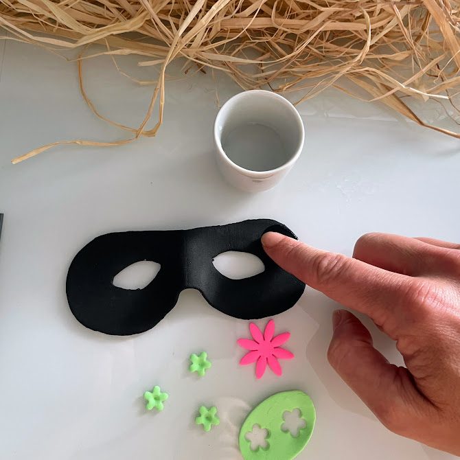Masque noir sur la table, doigt humidifiant une zone avant de coller des fleurs, récipient d’eau en haut.