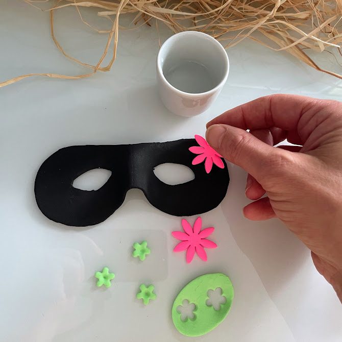 Main appliquant une grande fleur rose sur le masque, d'autres fleurs prêtes à être ajoutées.